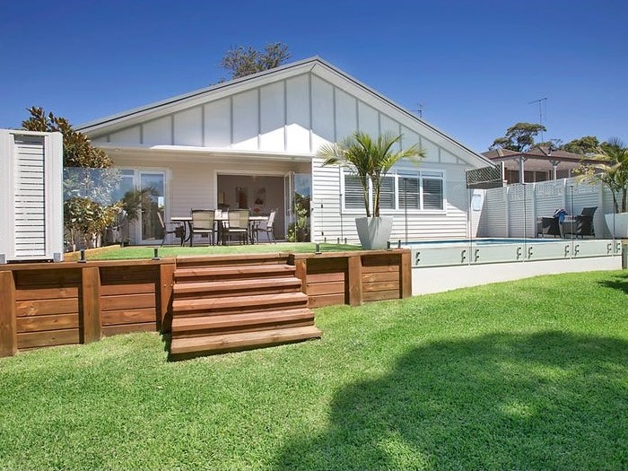 White coastal house with timber stairs leading up to back deck. Green lawn